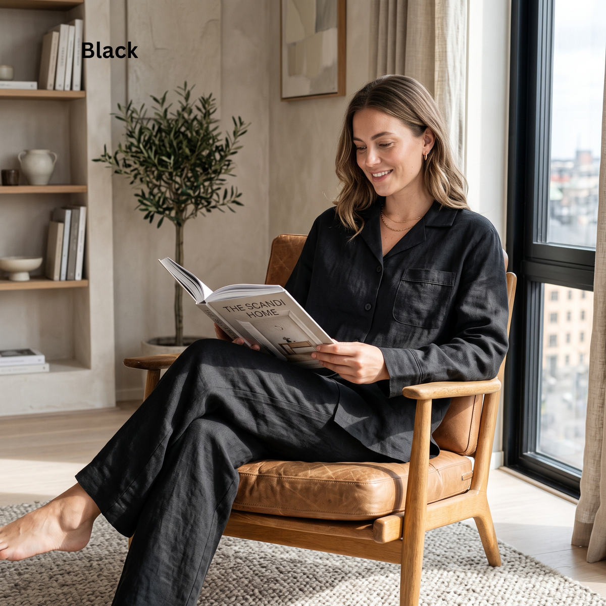A woman sits in a wooden armchair by a window, reading a book while wearing a black pajama set. The image includes the text 'Black' in the upper left corner, indicating the color option.