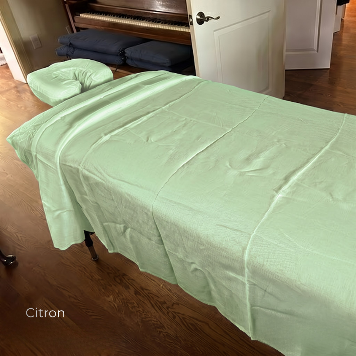 A light green massage table cover draped over a table in a home setting, with a matching headrest pillow. The room has hardwood floors and a white door in the background.