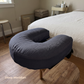 A dark gray U-shaped neck pillow resting on a wooden stool in front of a bed with white bedding in a softly lit bedroom. The text 'Deep Meridian' appears in the lower-left corner.