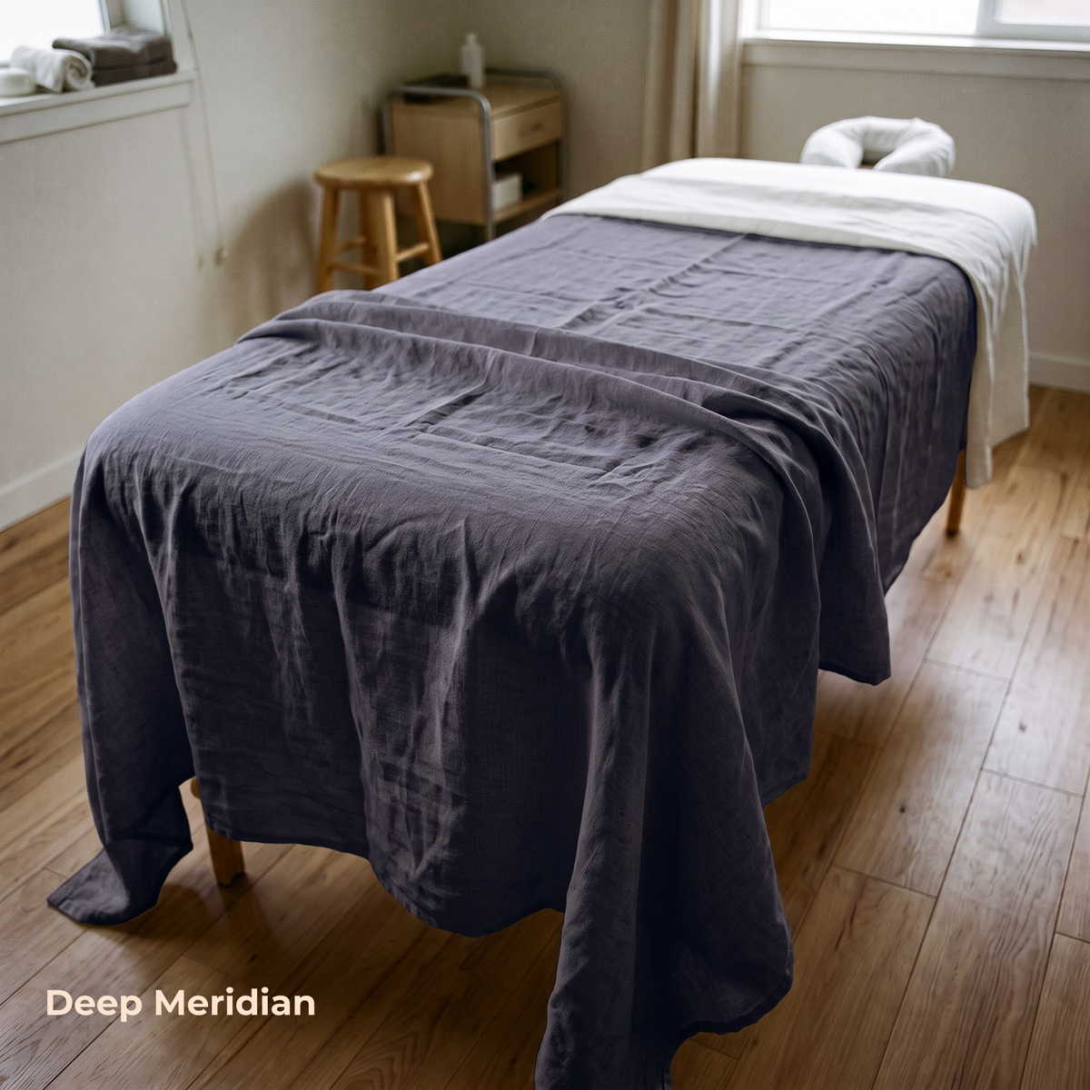 A massage table draped in a dark charcoal gray linen sheet, with a white towel and headrest on top, set in a bright room with wooden floors and a window. The text 'Deep Meridian' is visible in the bottom-left corner.