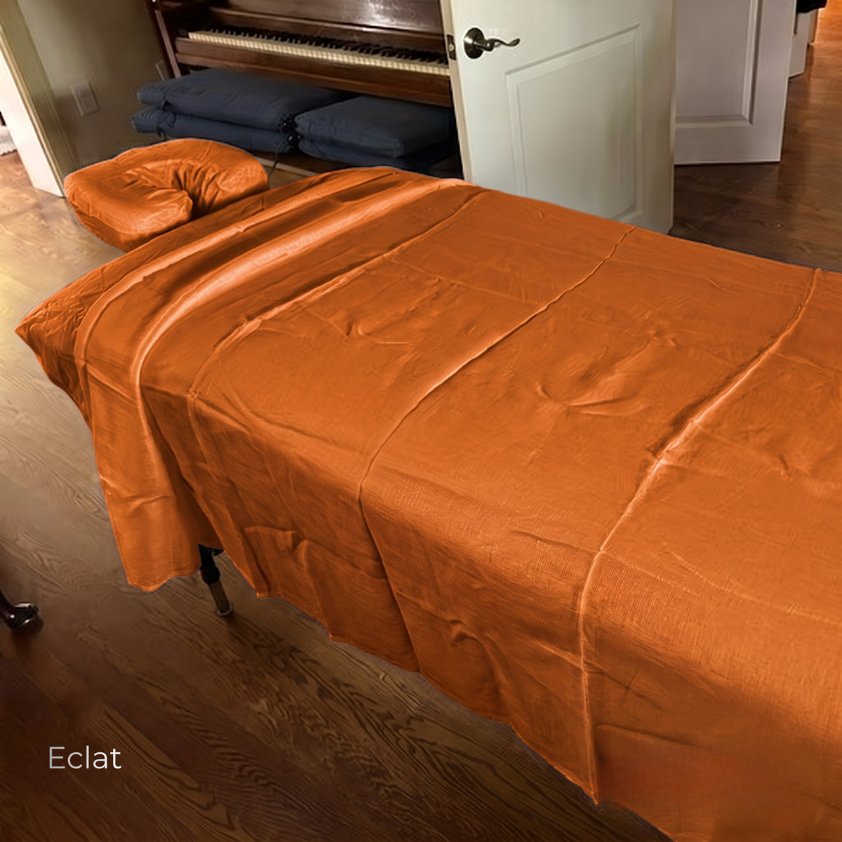 An orange satin bedspread and matching pillowcase draped over a massage table in a home office setting with a wooden floor and piano visible in the background.
