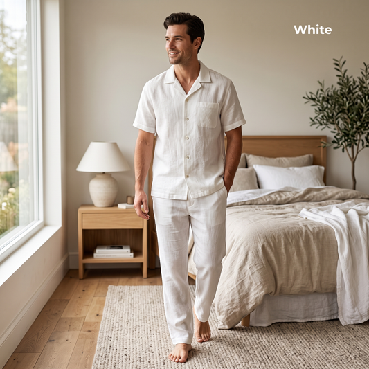 A man wearing a white short-sleeve button-front pajama shirt and matching pajama pants walks barefoot in a softly lit bedroom. The image includes the word 'White' in the top right corner.