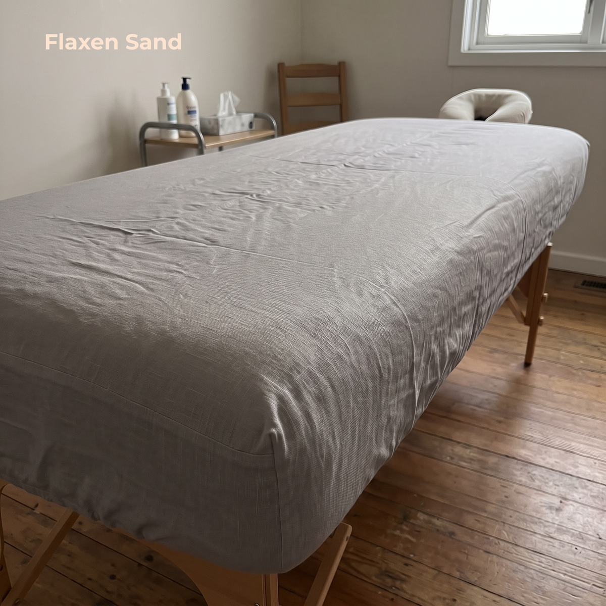 A light gray textured sheet or duvet cover spread across a wooden massage table in a minimalist room with hardwood floors, a window, and a small side table in the background.