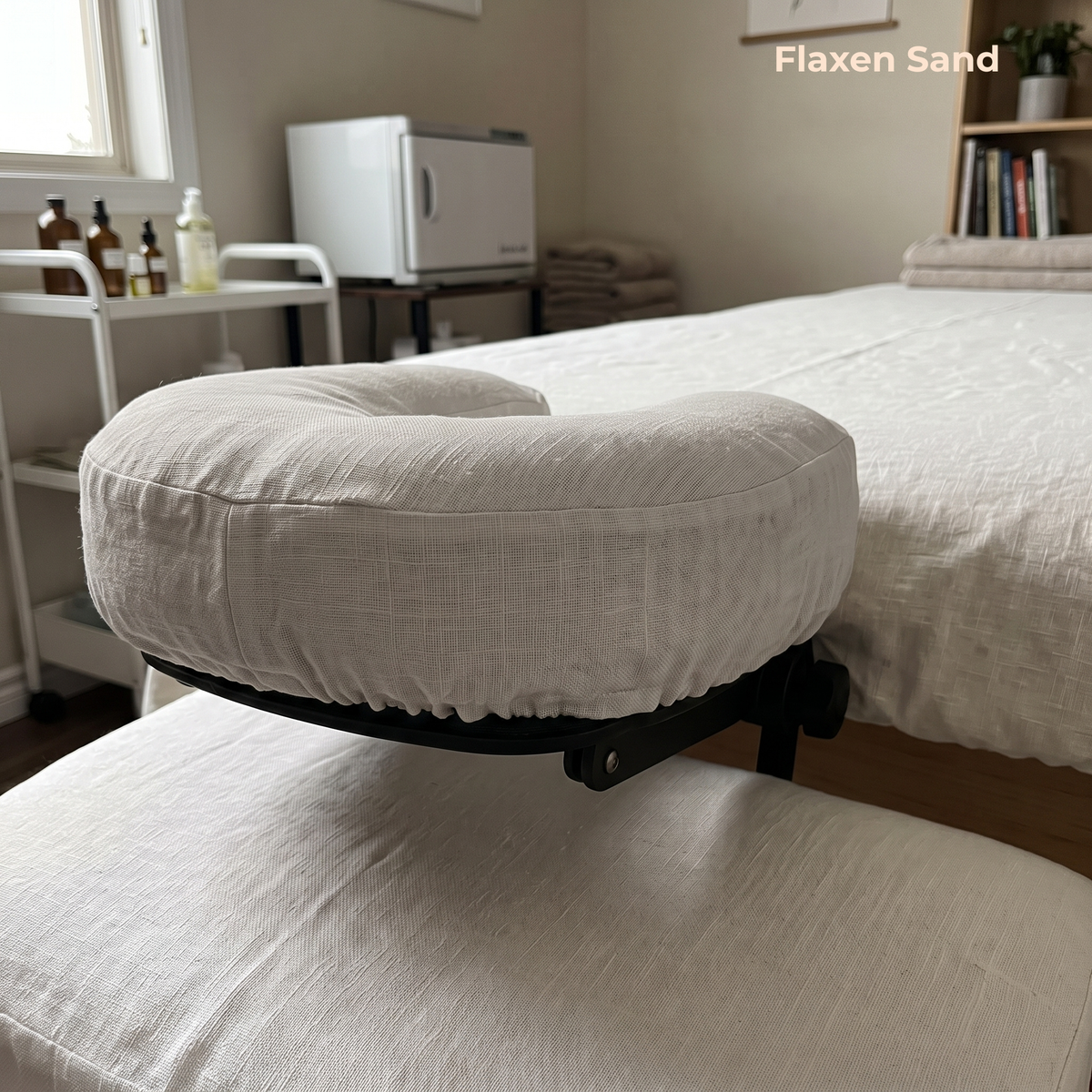 A close-up of a massage table headrest cushion upholstered in a textured off-white fabric, labeled 'Flaxen Sand,' set in a clean, minimalist spa room with a treatment bed and small accessories in the background.
