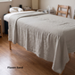 A massage table covered with a light beige linen sheet in a softly lit room with wood floors. The text 'Flaxen Sand' appears in the lower-left corner.