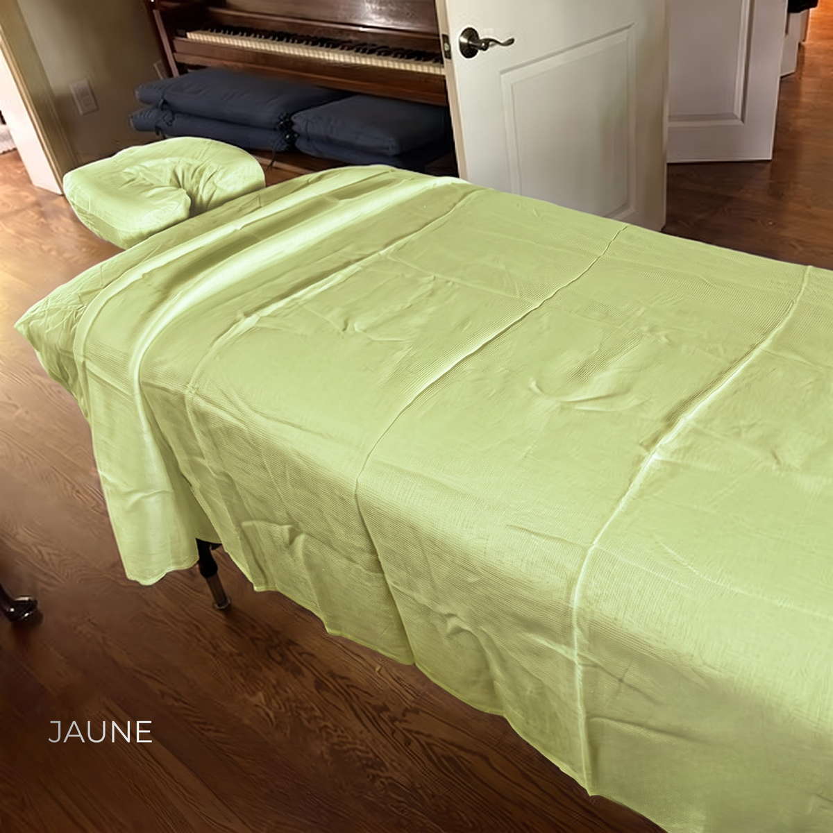 A light yellow-green massage table cover and matching headrest pad displayed on a massage table in a home setting with hardwood floors and a piano in the background. The word 'JAUNE' is printed in white in the bottom left corner.
