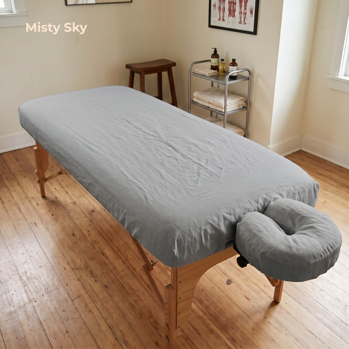 A gray massage table cover fitted over a wooden massage table in a spa-like room with wood floors, a small stool, and a metal cart. A matching gray headrest cushion is attached at the front.
