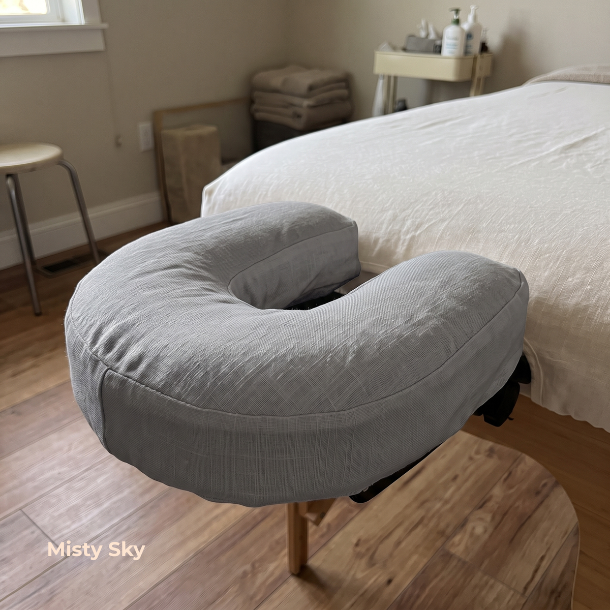 A light gray U-shaped massage pillow resting on a wooden table in front of a bed in a softly lit bedroom. The text 'Misty Sky' is visible in the bottom-left corner.