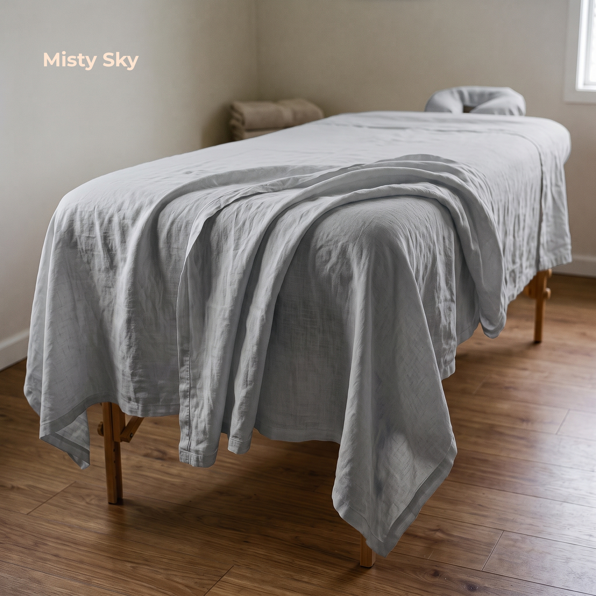 A light gray linen sheet set draped over a wooden massage table in a minimalist room with beige walls and a wood floor. The words 'Misty Sky' are printed in the upper left corner.