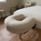 A beige U-shaped massage pillow labeled 'Oat Dune' resting on the head of a bed in a softly lit bedroom or spa-like room with wood floors and neutral walls.
