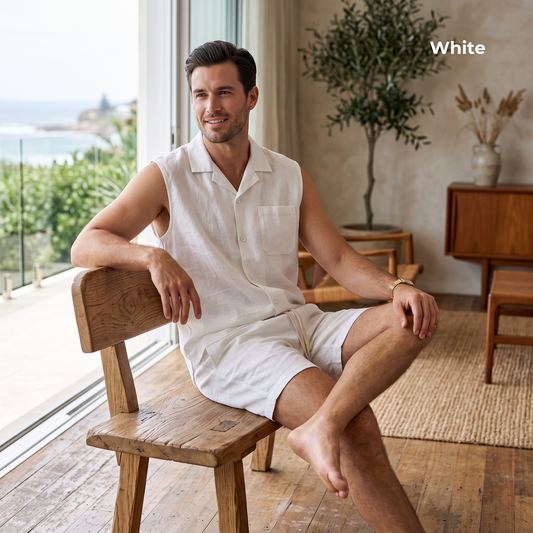 A man sits barefoot on a wooden chair in a bright, modern room, wearing a white sleeveless button-up shirt and matching white shorts. The word 'White' appears in the top right corner.