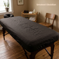 A dark charcoal gray massage table cover fitted on a massage table in a spa-like room with wooden floors, a wooden chair, and a small wooden shelf with towels and bottles.
