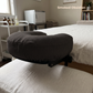 A padded massage or spa support cushion in a dark smoked obsidian fabric, resting on a white treatment table in a softly lit bedroom or spa room. The words 'Smoked Obsidian' appear in the upper right.