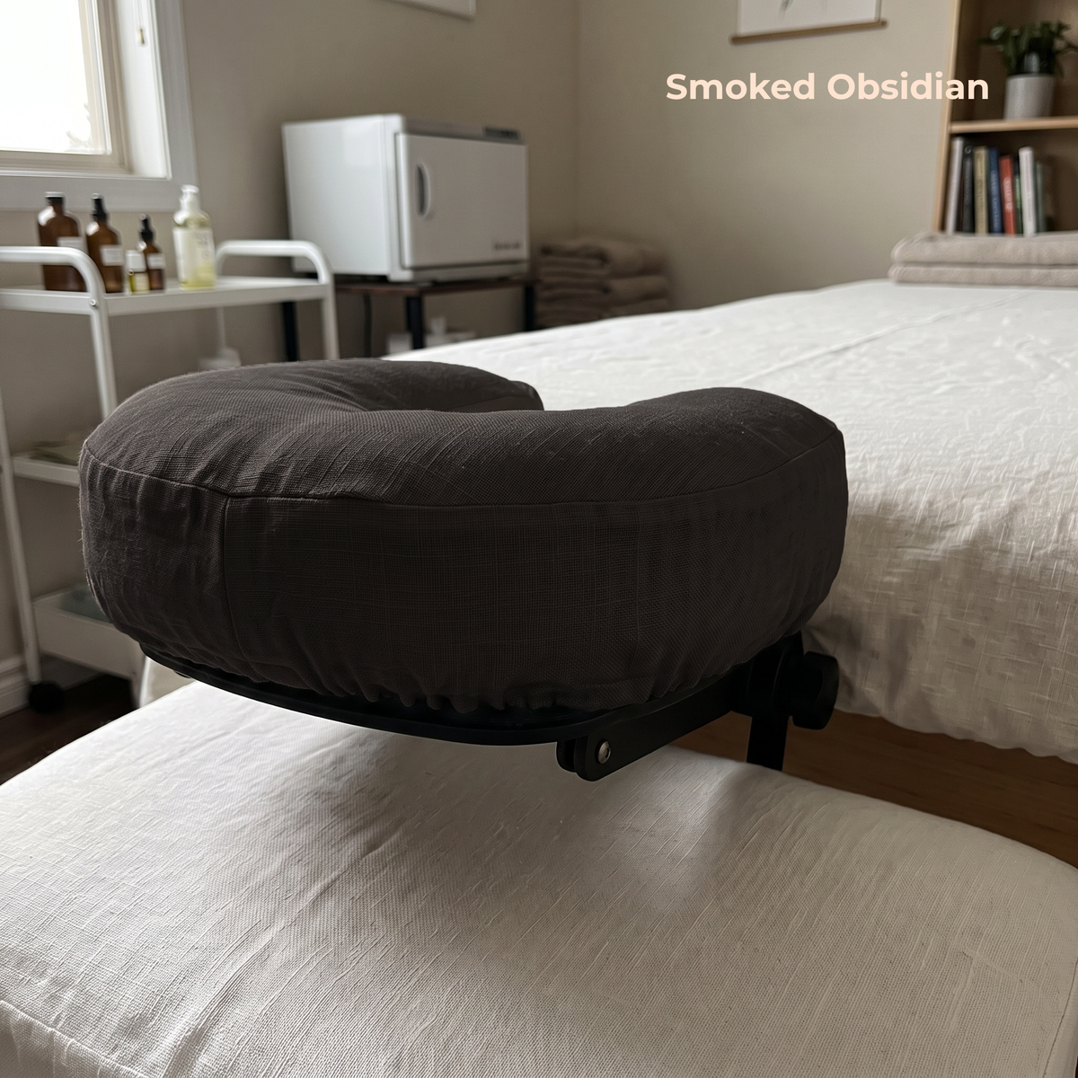 A padded massage or spa support cushion in a dark smoked obsidian fabric, resting on a white treatment table in a softly lit bedroom or spa room. The words 'Smoked Obsidian' appear in the upper right.