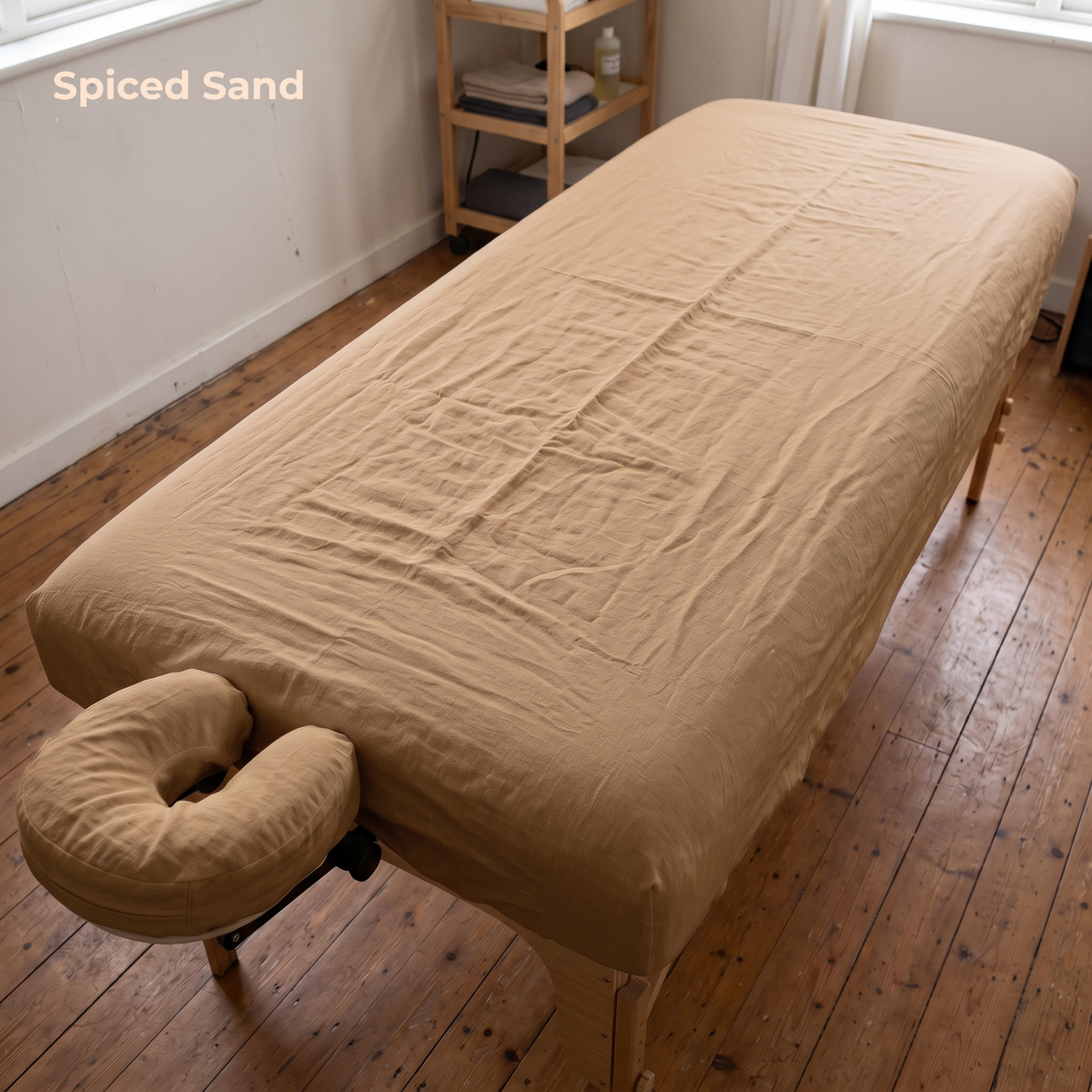 A beige massage table cover with an integrated U-shaped neck pillow, shown on a wooden floor in a room with a wooden shelf in the background. The text 'Spiced Sand' is visible in the top left corner.