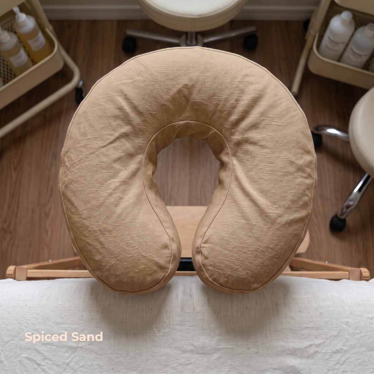 A U-shaped massage pillow in a warm beige fabric, displayed on a wooden table over a white sheet in what appears to be a spa or treatment room. The text 'Spiced Sand' is visible in the bottom-left corner.