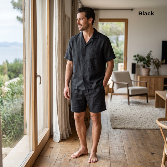 A man wearing a matching black short-sleeve button-up shirt and shorts pajama set stands barefoot in a bright, modern room with large windows overlooking a garden.