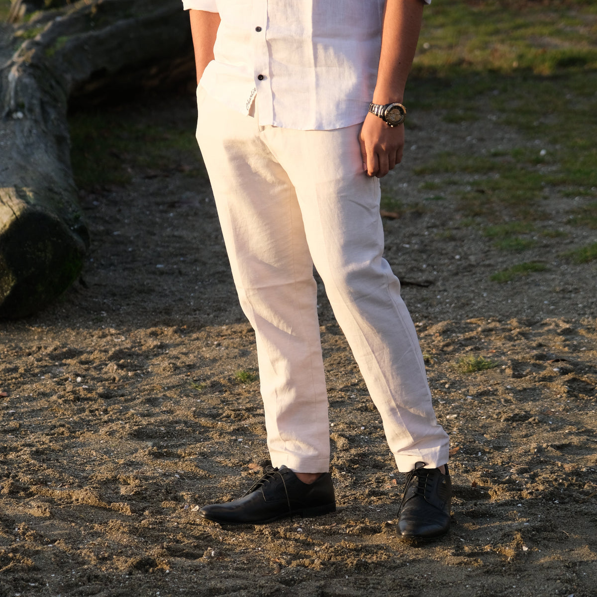 A person outdoors wearing light beige linen trousers, a white short-sleeve button-up shirt, and black shoes, standing on sandy ground with a log in the background.