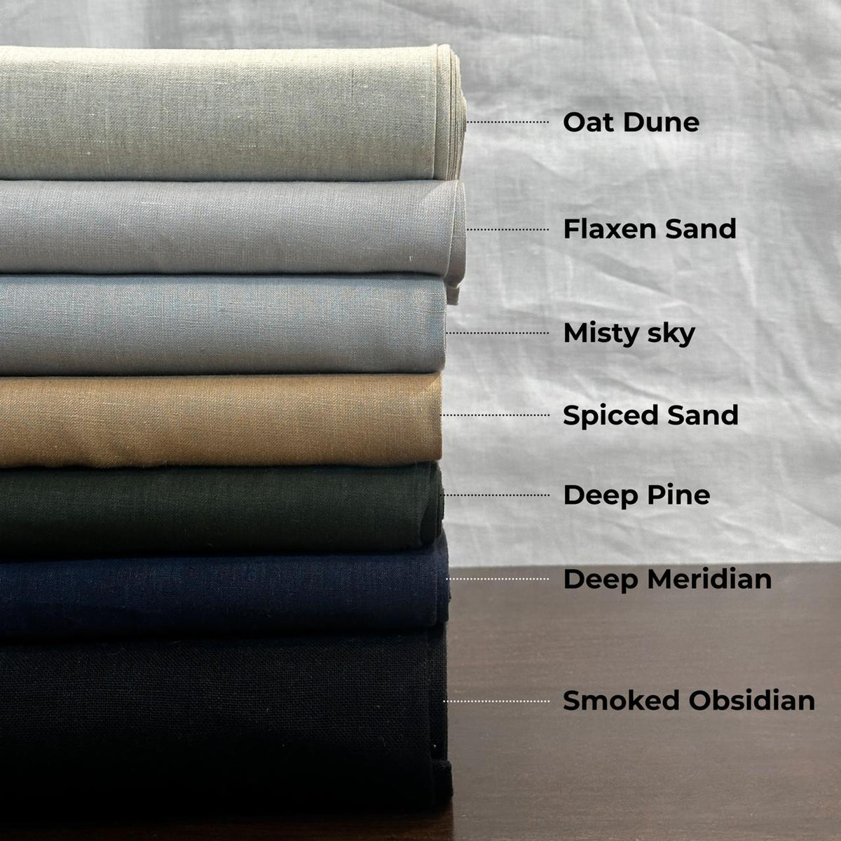 Six folded fabric swatches are stacked vertically on a white sheet, with text labels pointing to each color: Oat Dune, Flaxen Sand, Misty sky, Spiced Sand, Deep Pine, Deep Meridian, and Smoked Obsidian.